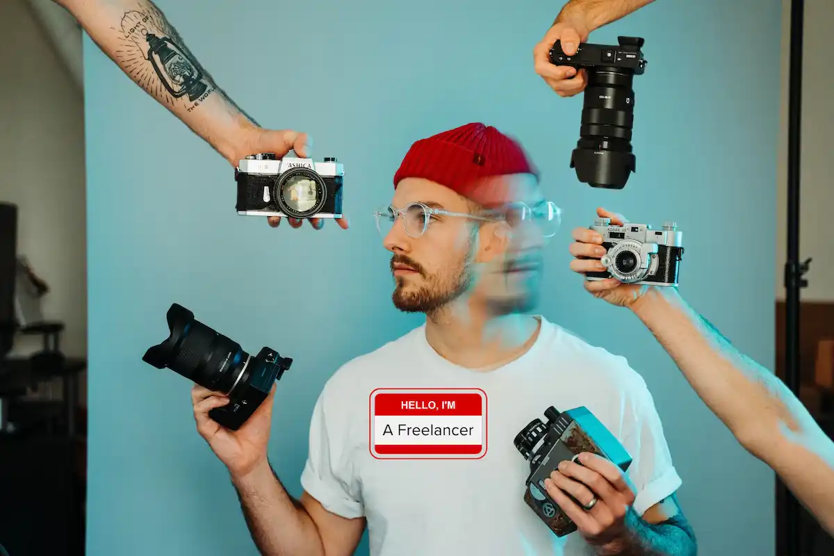 Man holding 2 cameras surrounded by other cameras, wearing a name badge that says he's a freelancer.